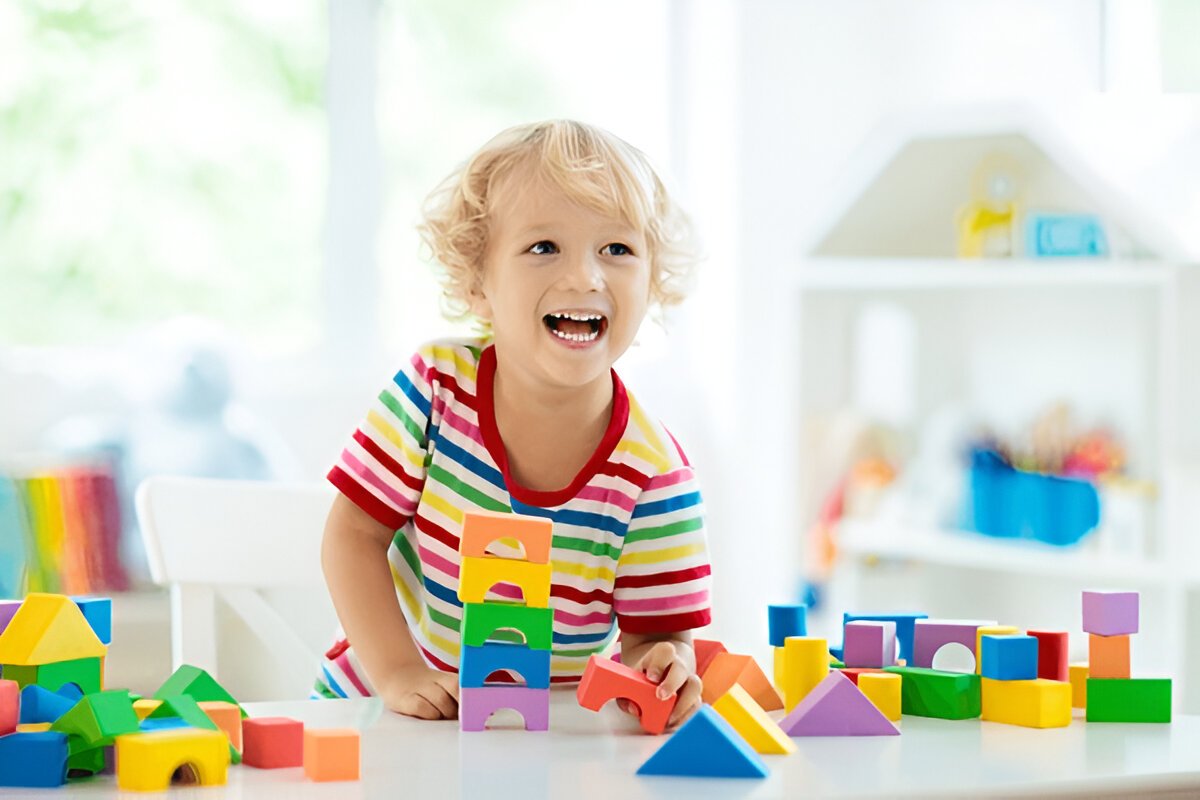 Smiling toddler playing with colorful building blocks in a bright preschool classroom.
