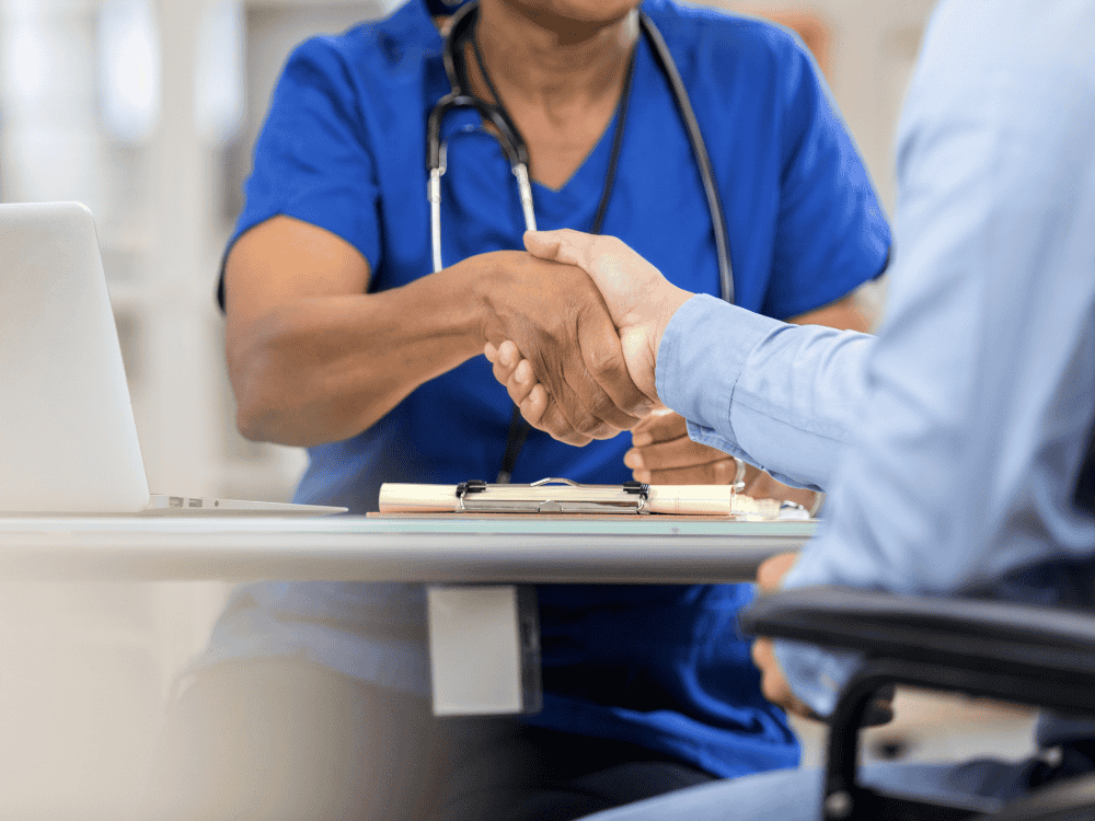 A dentist in blue scrubs shaking hands with a patient across a desk, symbolizing trust and partnership in dental care.
